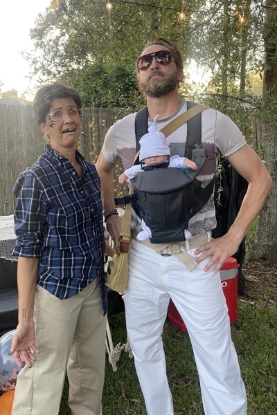 adult couple pose outdoors, dressed in costumes. The person on the left has face paint, wears a blue plaid shirt, khaki pants, and has a humorous, shocked expression. The guy on the right, wearing sunglasses and a beard, has a baby doll strapped to their chest in a baby carrier, dressed casually in a graphic tee and white pants.